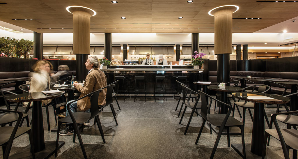 Interno elegante di Peck Milano: tavoli, sedie, bancone bar e persone. Design raffinato, atmosfera accogliente.