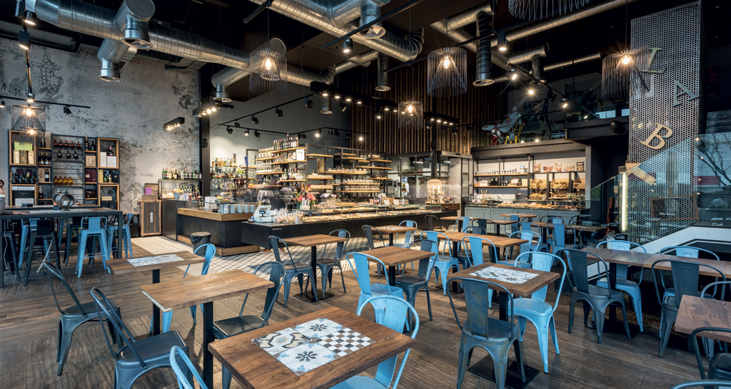 Interno di panetteria/caffè. Tavoli in legno, sedie blu, illuminazione industriale. Vetrina con prodotti da forno.