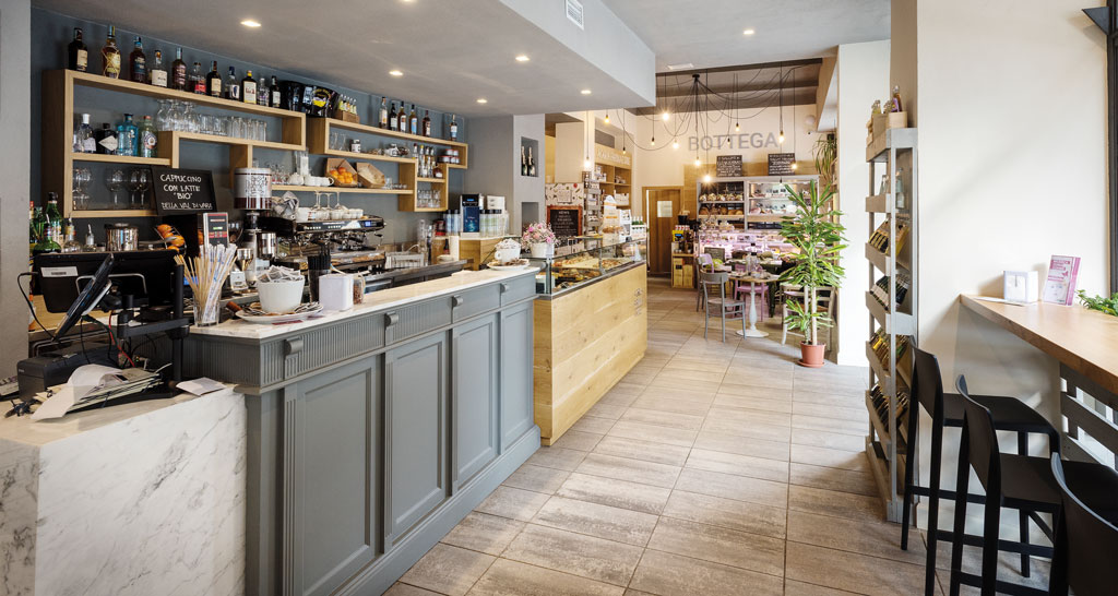 Interno di bottega italiana: bancone bar, scaffali con prodotti tipici e tavoli.