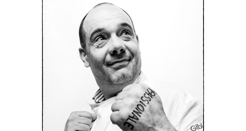 Ritratto in bianco e nero di chef sorridente con tatuaggio Passionale sulla mano. Cucina italiana.