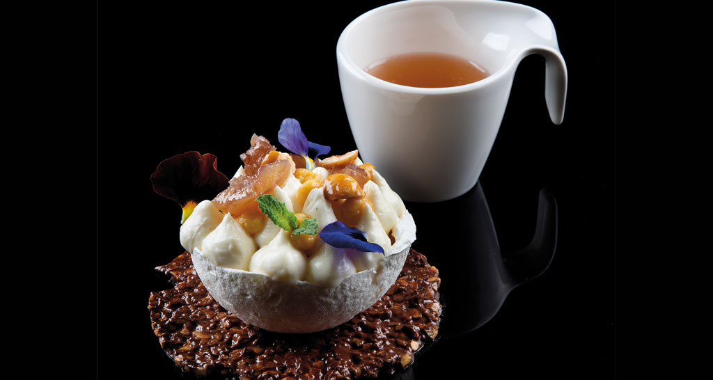 Dolce gourmet con crema, frutta e fiori, accanto a una tazza di tè. Dessert elegante su fondo nero.