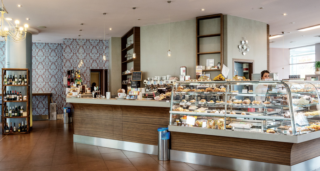 Interno di caffetteria: bancone con dolci, scaffale vini, illuminazione elegante. Arredamento accogliente.