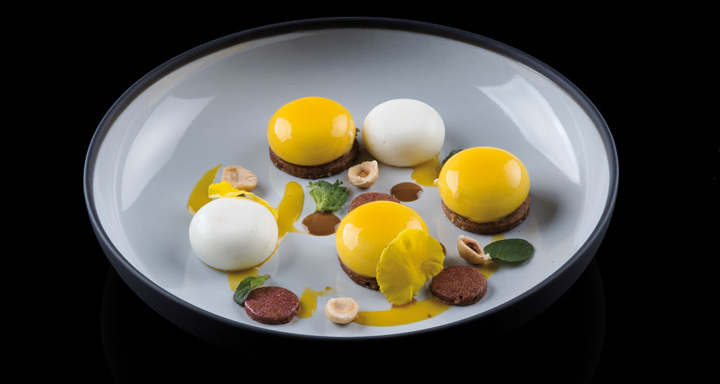 Dolci gourmet: dessert con sfere gialle e bianche, nocciole e decorazioni. Piatto elegante, alta cucina.