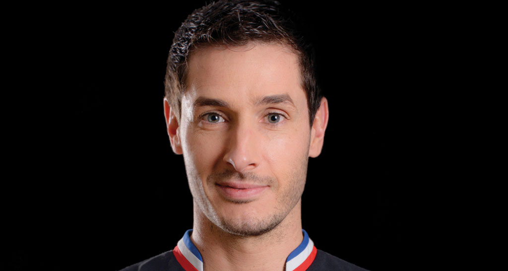 Ritratto frontale di chef uomo caucasico con divisa tricolore. Sguardo diretto, sfondo nero.