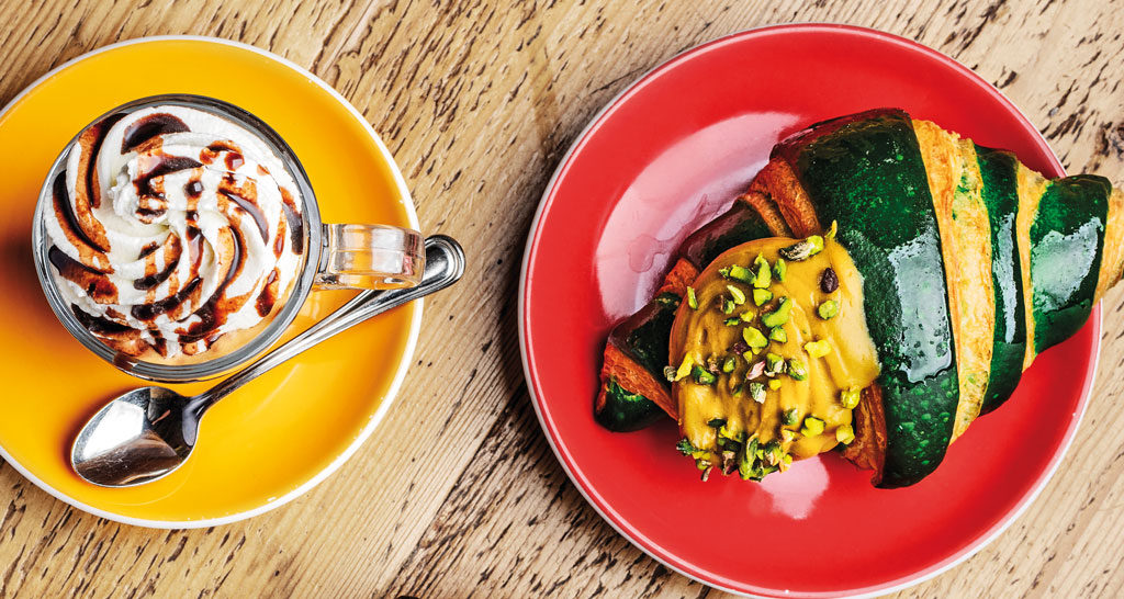 Colazione: cappuccino con panna e cioccolato, croissant al pistacchio su piatti colorati.