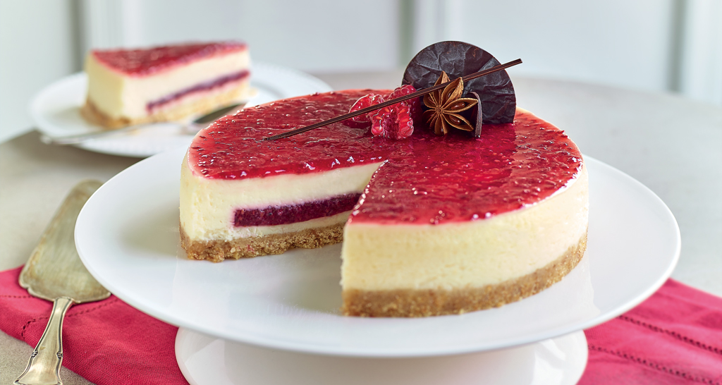 Cheesecake ai lamponi su alzata bianca. Dessert goloso con glassa rossa e decorazioni di cioccolato.