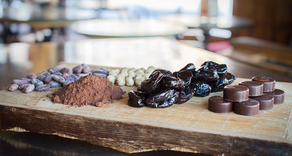 Prugne della California, cioccolato, fave di cacao, e cioccolatini artigianali su tagliere di legno.