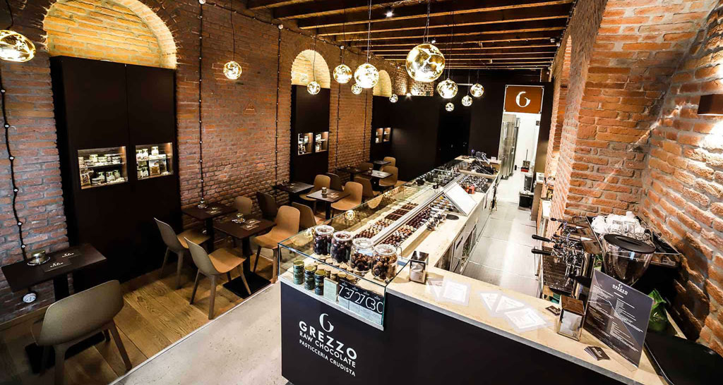 Interno di Grezzo Raw Chocolate: pasticceria, tavolini, luci soffuse, mattoni a vista. Design accogliente.