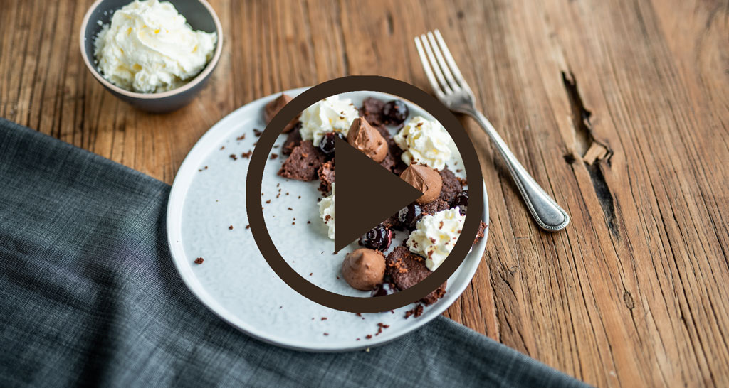 Dolce Foresta Nera con panna, cioccolato e ciliegie su piatto. Forchetta e sfondo in legno rustico.