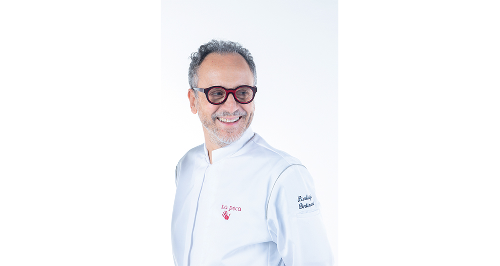 Chef Pierluigi Portinari sorridente. Divisa bianca, occhiali bordeaux. Cucina italiana, ristorante La Peca, alta cucina.