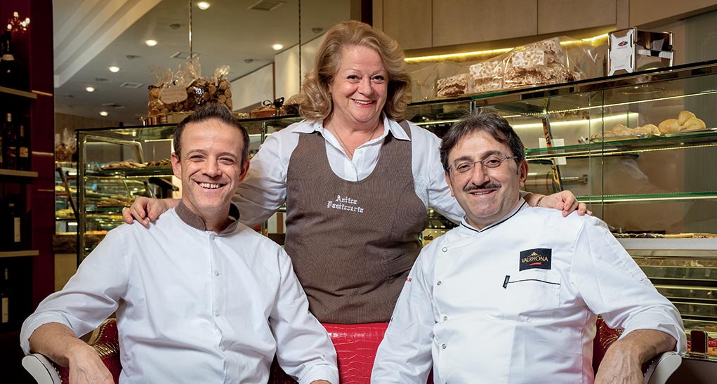 Pasticceria Artigianale Forlì: sorridenti pasticceri e proprietaria. Dolci e torte in vetrina sullo sfondo.