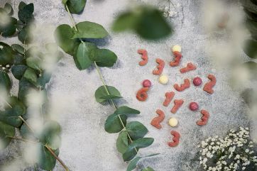 Biscotti frolla a forma di numeri e caramelle su sfondo grigio con eucalipto e fiori bianchi.