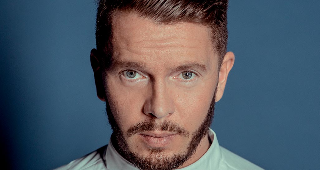 Ritratto uomo barbuto con occhi azzurri. Primo piano volto maschile, capelli castani. Espressione seria, sfondo blu.