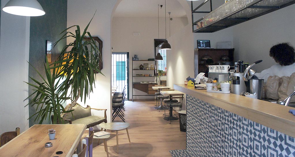 Interni del caffè Ofelé, Milano. Design moderno con piante, tavoli in legno e bancone decorato. Atmosfera accogliente.