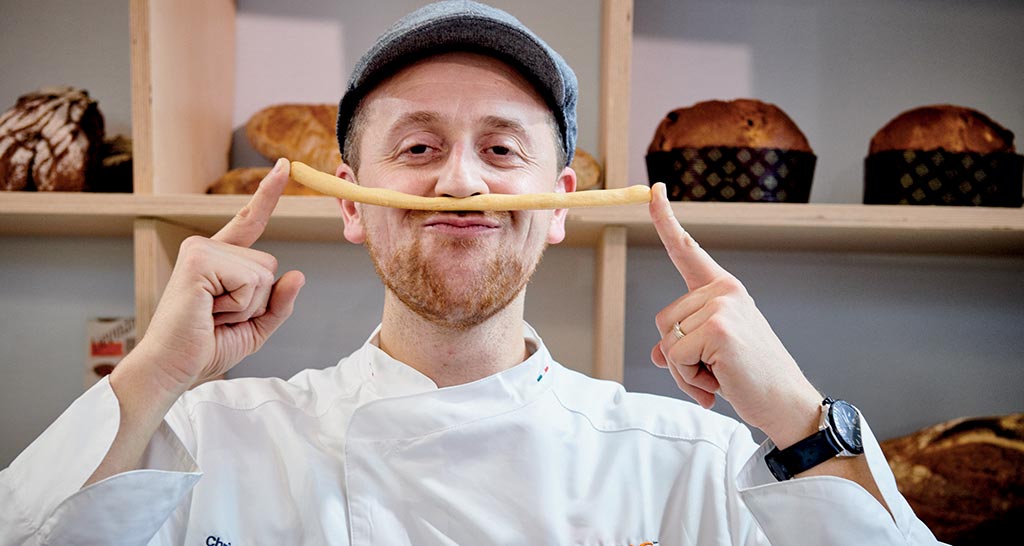 Chef panettiere sorridente con grissino. Pane e dolci artigianali sullo sfondo. #panetteria #chef #grissini