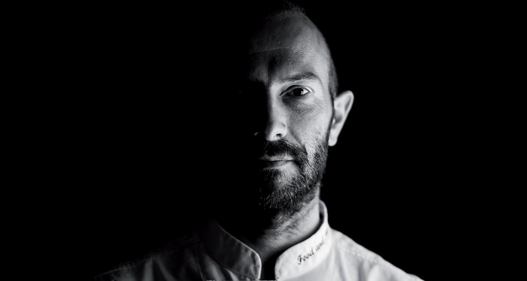 Ritratto in bianco e nero di Luca Bernardini, chef con barba, su sfondo nero. Cucina italiana.