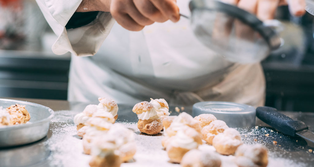 Pasticcere spolvera zucchero a velo su bignè con crema. Dolci artigianali.