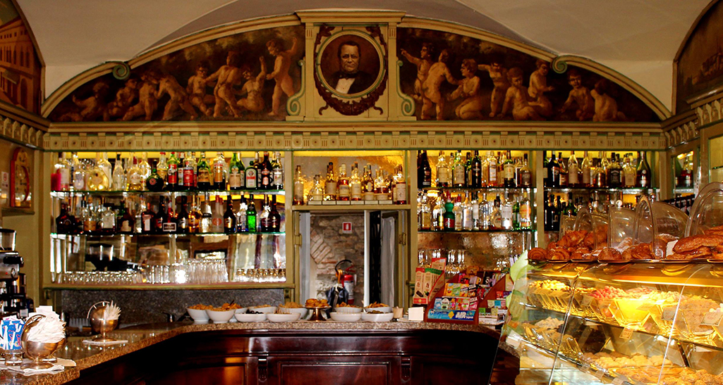 Interno bar storico: scaffali di liquori, dolci, affresco con Cavour.