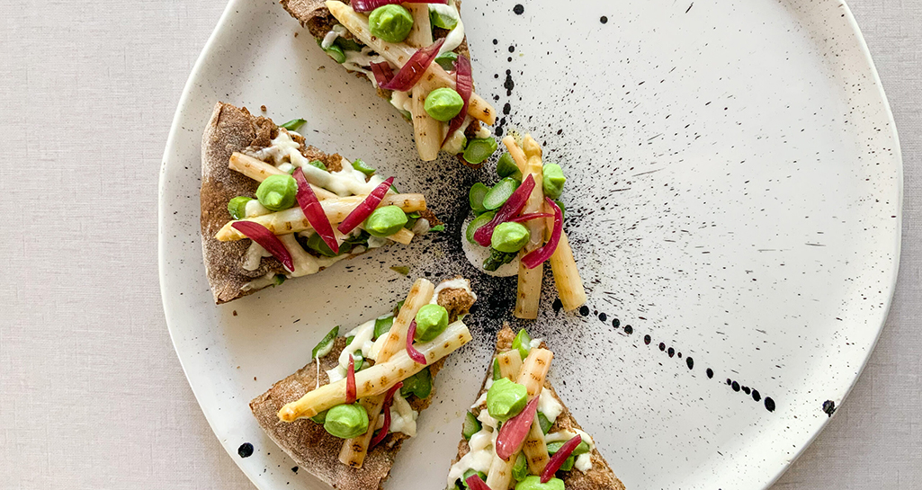Pizza gourmet con asparagi, fave e cipolle rosse caramellate su piatto decorato. Ricetta primaverile.