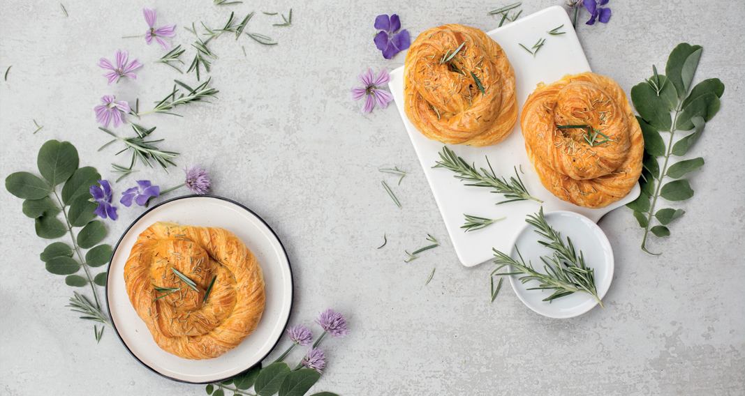Tre brioche artigianali con rosmarino e fiori viola. Dolci da forno su piatti bianchi.