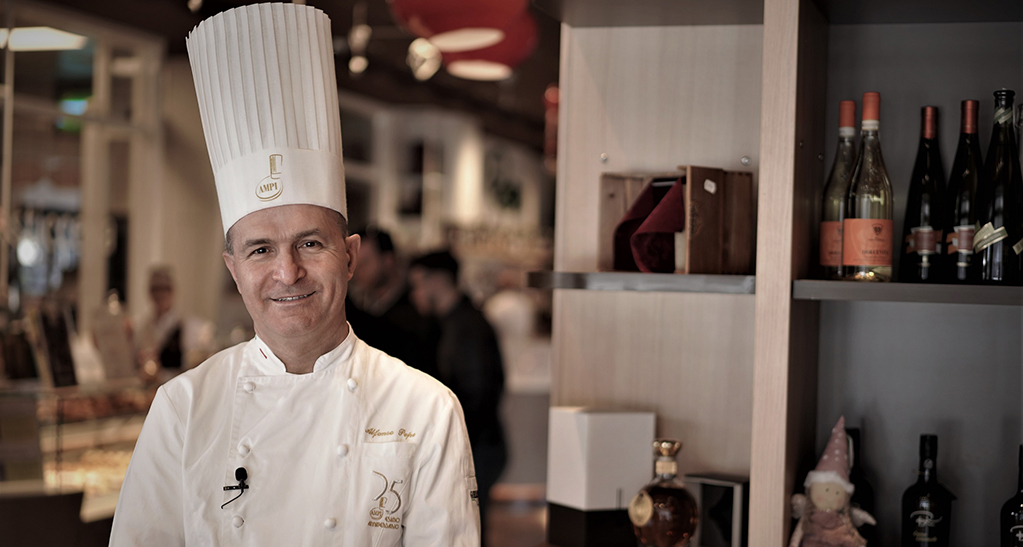 Chef Alfonso Pepe sorridente in divisa. Bottiglie di vino sullo sfondo.