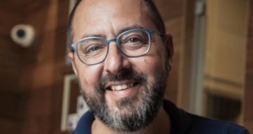 Ritratto di Alberto Marchetti, uomo sorridente con barba e occhiali azzurri.