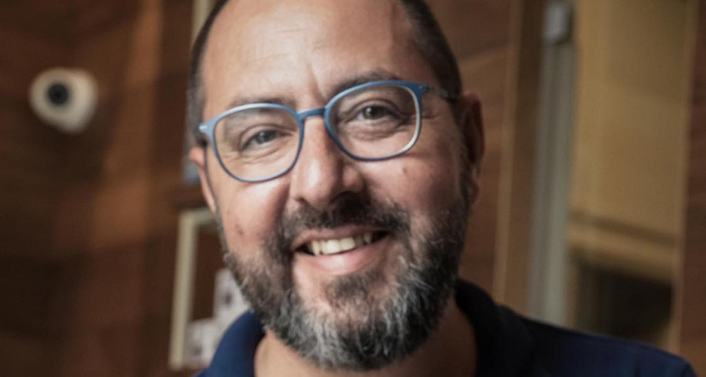 Ritratto di Alberto Marchetti, uomo sorridente con barba e occhiali azzurri.