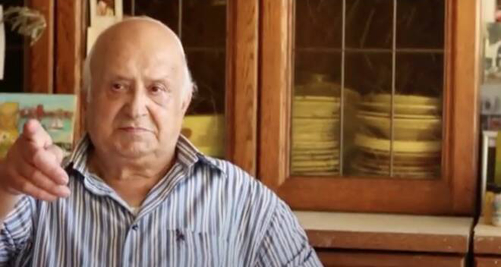 Ritratto di Carmine Marzuillo, artista italiano. Uomo calvo con camicia a righe indica qualcosa. Arte, cultura italiana.