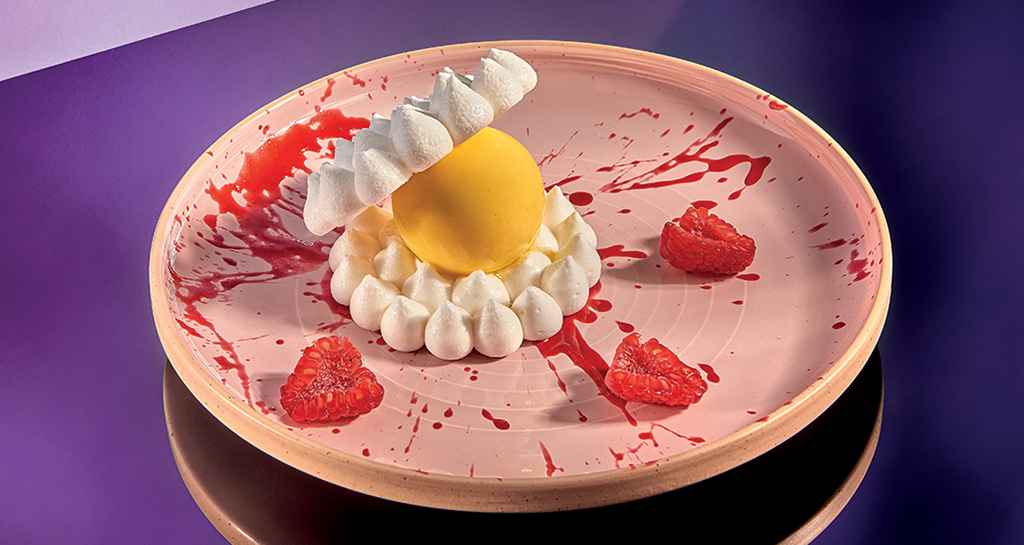 Dolce esotico Perla Tropicale: sfera gialla, meringhe, lamponi su piatto rosa con decorazioni rosse. Dessert creativo.