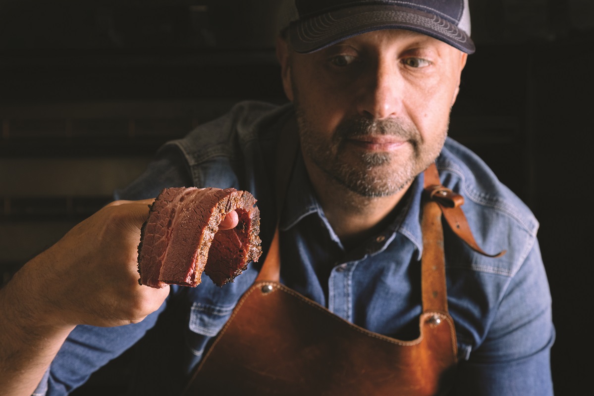 Joe Bastianich con carne. Chef americano con grembiule di cuoio mostra un pezzo di carne. Cucina USA autentica.