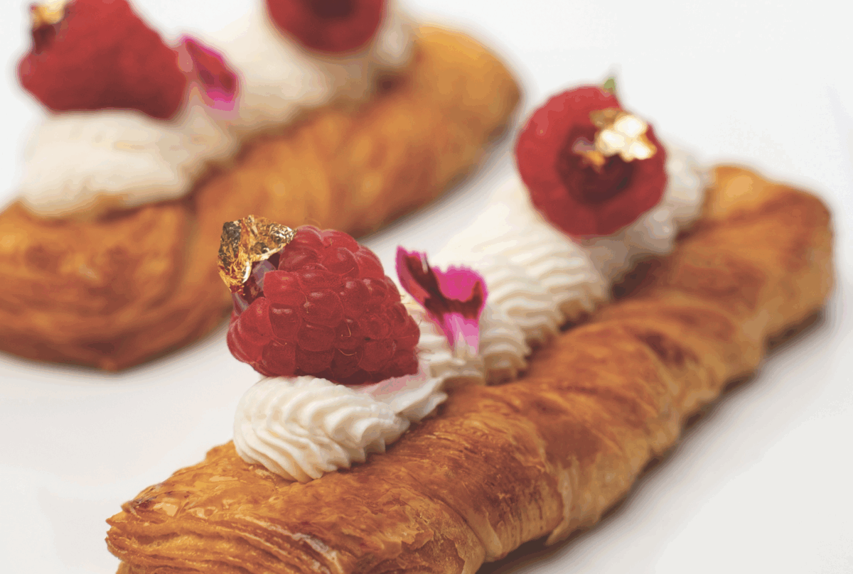 Treccia sfogliata senza lattosio guarnita con crema bianca, lamponi freschi e decorazioni dorate su sfondo bianco.