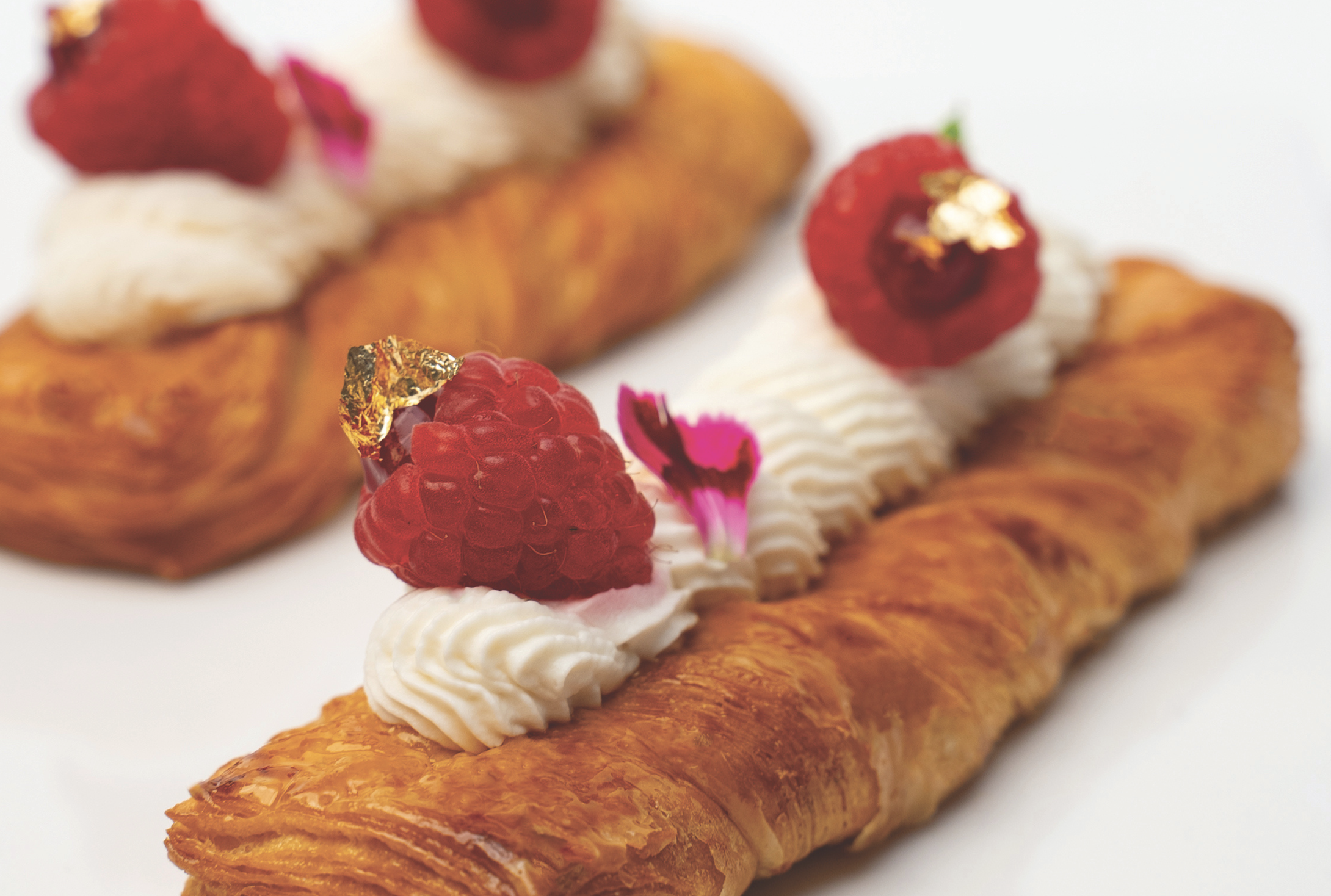 Treccia sfogliata senza lattosio guarnita con crema bianca, lamponi freschi e decorazioni dorate su sfondo bianco.
